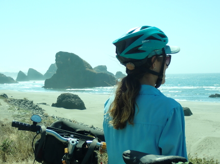 Ruth on the coast with bike in the foreground and rocks protruding out of the sea behind