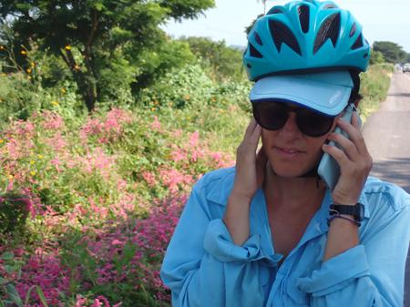 Ruth with her ear to her mobile on the roadside with pretty flowers in the background