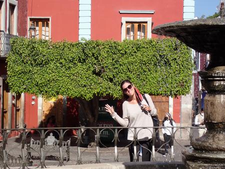 Ruth waving behind a flowing fountain in a Guanajuaton square with a pruned tree in the background