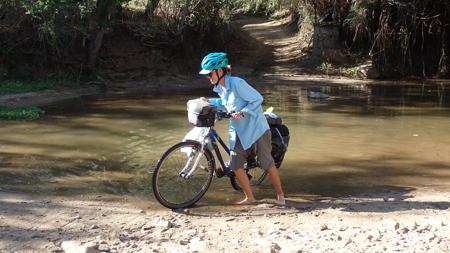Ruth coming out of a river barefoot after pushing her bike through the obstacle